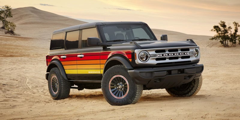 Black Ford Bronco with red, orange, and yellow retro sunset stripes on its side, parked on sand dunes.