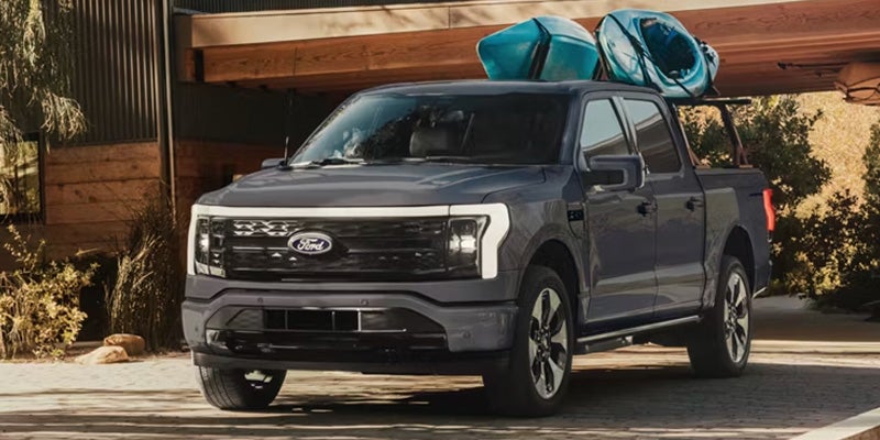 Dark gray Ford F-150 truck with two blue kayaks on a rack in its bed, parked in front of a modern house.